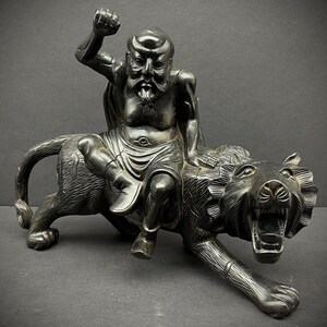 May include: A black wood carving of a man riding a tiger. The man is wearing a robe and has a beard. The tiger is snarling with its mouth open.