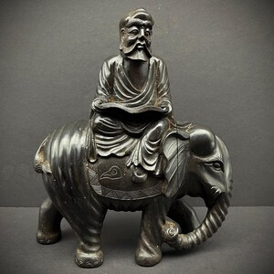 May include: A black wood carving of a man sitting on an elephant. The man is wearing a robe and has a beard. The elephant has a trunk and tusks.
