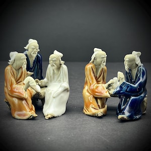 May include: Four ceramic figurines of elderly men in traditional Chinese clothing, sitting and interacting with each other. The figurines are painted in shades of blue, white, and brown.