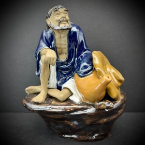 May include: A ceramic figurine of a man with a long beard, wearing a blue robe and sitting on a rock with a brown teapot. The figurine is hand-painted with details in brown, blue, and white.