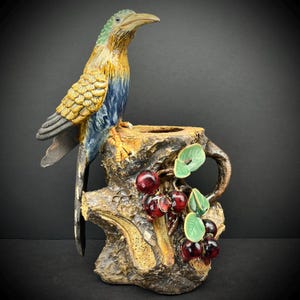 May include: A ceramic vase shaped like a tree stump with a bird perched on top. The vase is decorated with red berries and green leaves. The bird is blue, green, and gold.