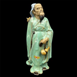 May include: A ceramic figurine of an elderly man in a long, flowing robe. The figurine is primarily light green with gold accents and a black beard. The man is holding two gourds and has a serene expression.