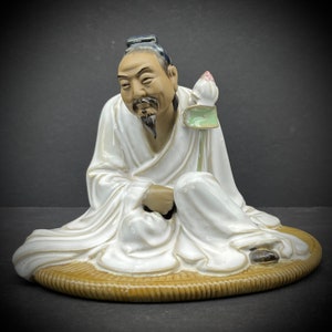 May include: A ceramic figurine of a seated man with a long white beard and a lotus flower in his hand. He is wearing a white robe and is sitting on a woven mat. The figurine is painted in shades of white, brown, and green.