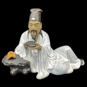 May include: A ceramic figurine of a man in a white robe and a black hat, sitting on a rock and holding a small dish. The figurine is painted in shades of brown, white, and black.