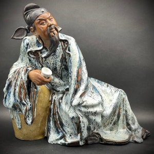 May include: A ceramic figurine of a man wearing a blue and white robe with a white cup in his hand. The figurine is sitting on a brown base.