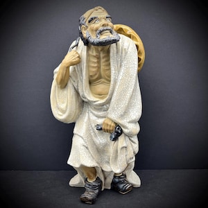 May include: A ceramic figurine of a man with a long white beard and a white robe. He is carrying a large, round object on his back. The figurine is standing on a base and has a cracked glaze finish.