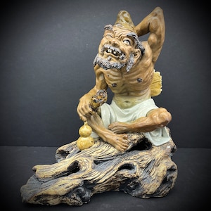 May include: A ceramic figurine of a mythical creature with a long beard and a gourd in its hand. The creature is sitting on a tree stump with a gnarled, textured surface.