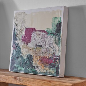 Op de afbeelding: Abstract schilderij op canvas met een wit wolfsilhouet in een groen, roze en gouden landschap. Het schilderij heeft een textuur en is ingelijst in een wit frame.