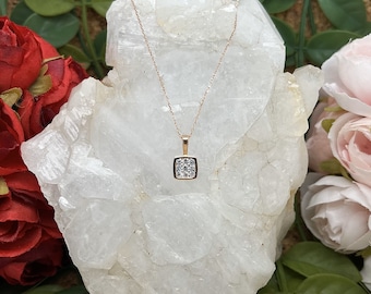 May include: A delicate gold necklace with a square pendant set with small, clear stones. The necklace is displayed on a white, textured surface.