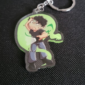 May include: A green and black cartoon character keychain with a silver key ring. The character is a person with dark hair and a green ghost behind them.