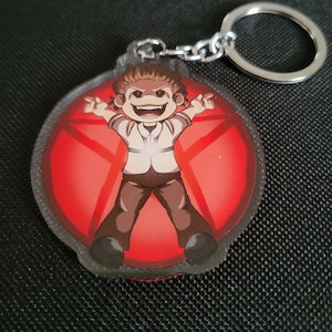 May include: A round, clear acrylic keychain with a red background and a cartoon character in the center. The character is a person with brown hair, a white shirt, and brown pants. The character is smiling and has red eyes. The character is standing with their arms outstretched. The background is a red circle with a black star in the center.