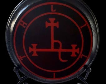 May include: Black round decorative plate with a red sigil design. The plate is displayed on a black metal stand. The sigil features a central cross design with the letters "L", "I", and "H" around the perimeter.