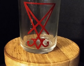 May include: A clear glass shot glass with a red inverted pentagram symbol.