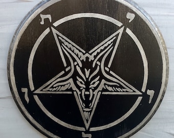 Puede incluir: Una placa circular de madera negra con un diseño de pentagrama grabado en color plateado. En el centro de la estrella hay una ilustración de una cabeza de cabra. Cinco caracteres hebreos están situados entre las puntas de la estrella.