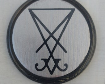 Puede incluir: Un símbolo en blanco y negro, conocido como el Sigil of Baphomet, se muestra sobre un fondo circular plateado con un borde negro.