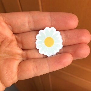 May include: A white daisy sticker with a yellow centre. The sticker is on a hand.