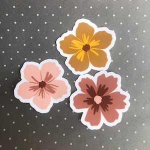 Puede incluir: Tres pegatinas de flores en diferentes colores. Las pegatinas son rosas, marrones y amarillas. Las flores son estilizadas y tienen un diseño simple.