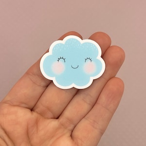 Puede incluir: Una pegatina de nube azul con una cara sonriente y mejillas rosadas. La nube tiene un contorno blanco y pequeños puntos blancos alrededor.