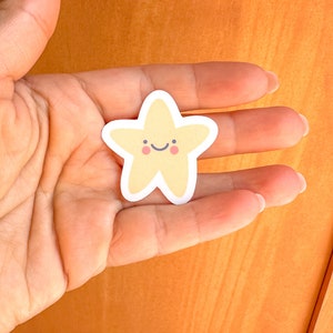 Puede incluir: Una pegatina amarilla en forma de estrella con una cara sonriente y mejillas rosadas. La pegatina está en una mano.
