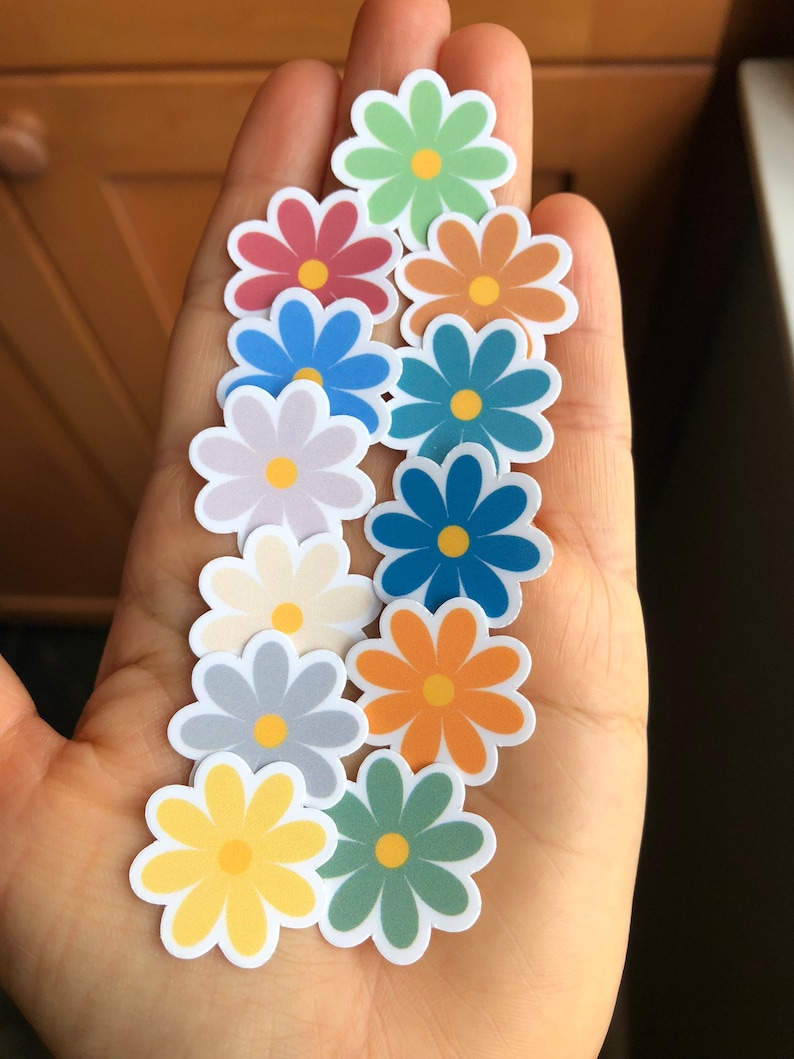 Puede incluir: Una colecci&oacute;n de 10 pegatinas de margaritas coloridas con centros amarillos. Las pegatinas est&aacute;n dispuestas en una mano, con la pegatina de margarita amarilla en el centro.