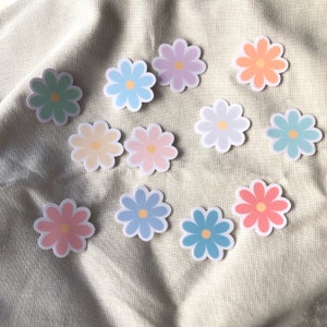 Puede incluir: Un conjunto de diez pegatinas de flores de margarita de color pastel. Las pegatinas son de varios tonos de azul, verde, rosa, amarillo y naranja.