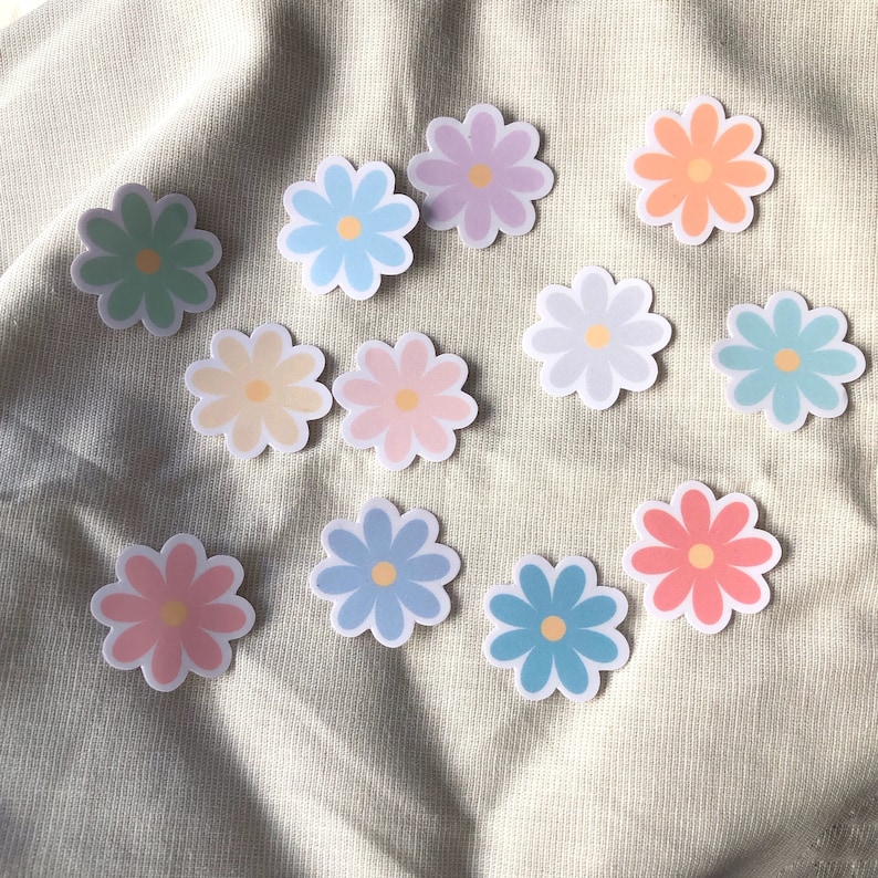 Puede incluir: Un conjunto de diez pegatinas de flores de margarita de color pastel. Las pegatinas son de varios tonos de azul, verde, rosa, amarillo y naranja.