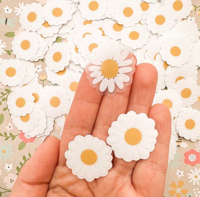 May include: A hand holding a variety of white daisy stickers with yellow centers. Some of the stickers are translucent and have a slightly raised texture.