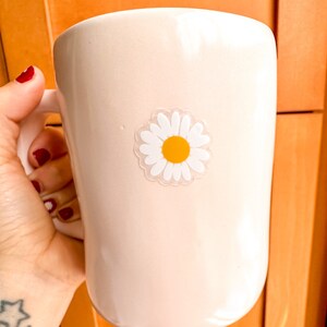 May include: A white ceramic mug with a daisy sticker on the side. The daisy has a yellow center and white petals.