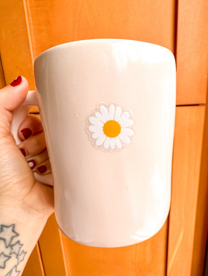 May include: A white ceramic mug with a daisy sticker on the side. The daisy has a yellow center and white petals.