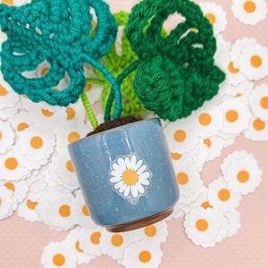 May include: A small crocheted plant with green leaves in a blue ceramic pot with a white daisy sticker. The pot is surrounded by white felt daisy shapes with orange centers.