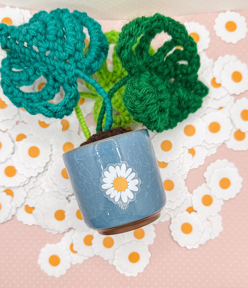 May include: A small crocheted plant with green leaves in a blue ceramic pot with a white daisy sticker. The pot is surrounded by white felt daisy shapes with orange centers.