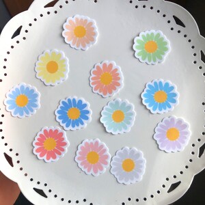 May include: A set of 12 pastel daisy stickers on a white plate with a scalloped edge. The stickers are in various pastel colours, including pink, blue, yellow, and green.