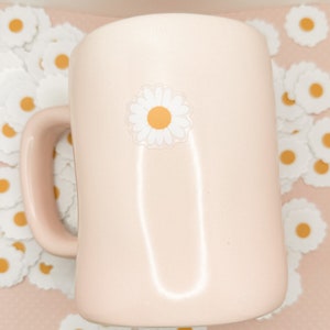 May include: A pink ceramic mug with a white daisy sticker on the front. The mug is surrounded by white daisy stickers on a pink background.