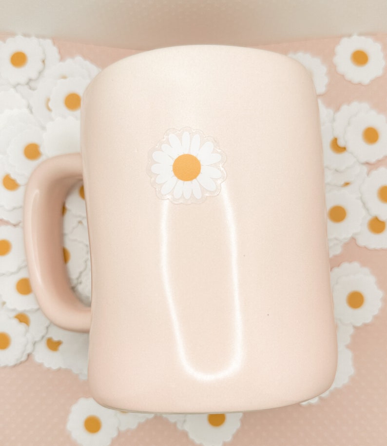 May include: A pink ceramic mug with a white daisy sticker on the front. The mug is surrounded by white daisy stickers on a pink background.