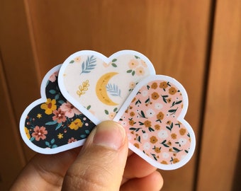 May include: Three heart-shaped stickers with different floral patterns. The top sticker has a cream background with blue leaves, a yellow crescent moon, and small pink flowers. The middle sticker has a black background with pink and yellow flowers. The bottom sticker has a pink background with small brown flowers and green leaves.