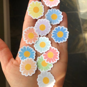 May include: A collection of pastel-coloured daisy stickers with yellow centres. The stickers are arranged on a hand.