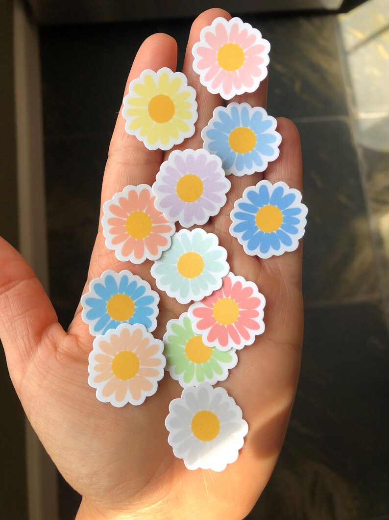 May include: A collection of pastel-coloured daisy stickers with yellow centres. The stickers are arranged on a hand.