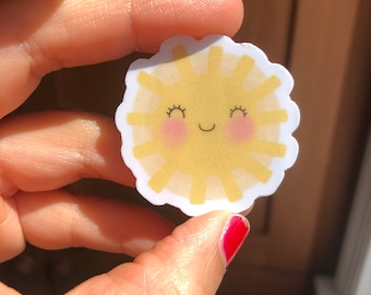 May include: A yellow sun sticker with a smiling face and closed eyes. The sun has a white outline and a white background.