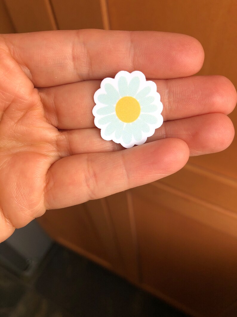May include: A white daisy sticker with a yellow centre. The sticker is on a hand.