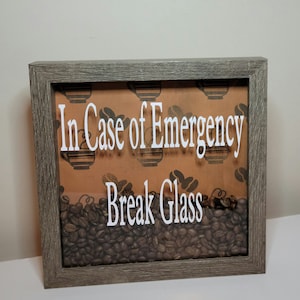 May include: A framed sign with a brown background and a white border. The sign says "In Case of Emergency Break Glass" and is filled with coffee beans.