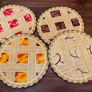 May include: Four round fabric coasters with a pie crust design. Each coaster features a different fruit filling: strawberry, peach, apple, and chocolate.