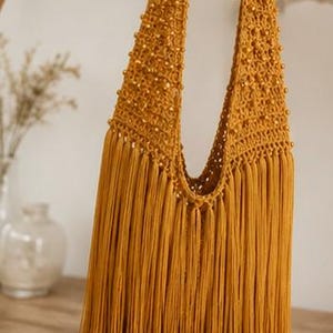 Puede incluir: Un bolso hobo de ganchillo color amarillo mostaza con un patrón de punto abierto adornado con pequeñas cuentas a juego en la correa y el cuerpo, terminado con un fleco largo y denso que cuelga de la base de la estructura del bolso.