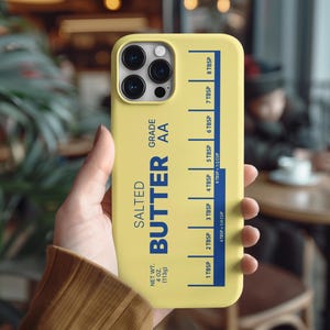 May include: A yellow phone case featuring a design inspired by a stick of butter. The case has the words "SALTED BUTTER AA" in blue, with measurement markings in tablespoons and cups. The net weight is 4 oz (113g).