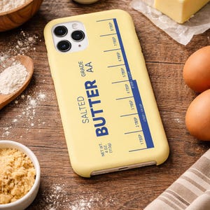 May include: A yellow phone case with a butter-themed design. The case features the words "SALTED BUTTER AA GRADE" and a measurement guide in tablespoons. Other baking ingredients like flour, eggs, and butter are in the background.