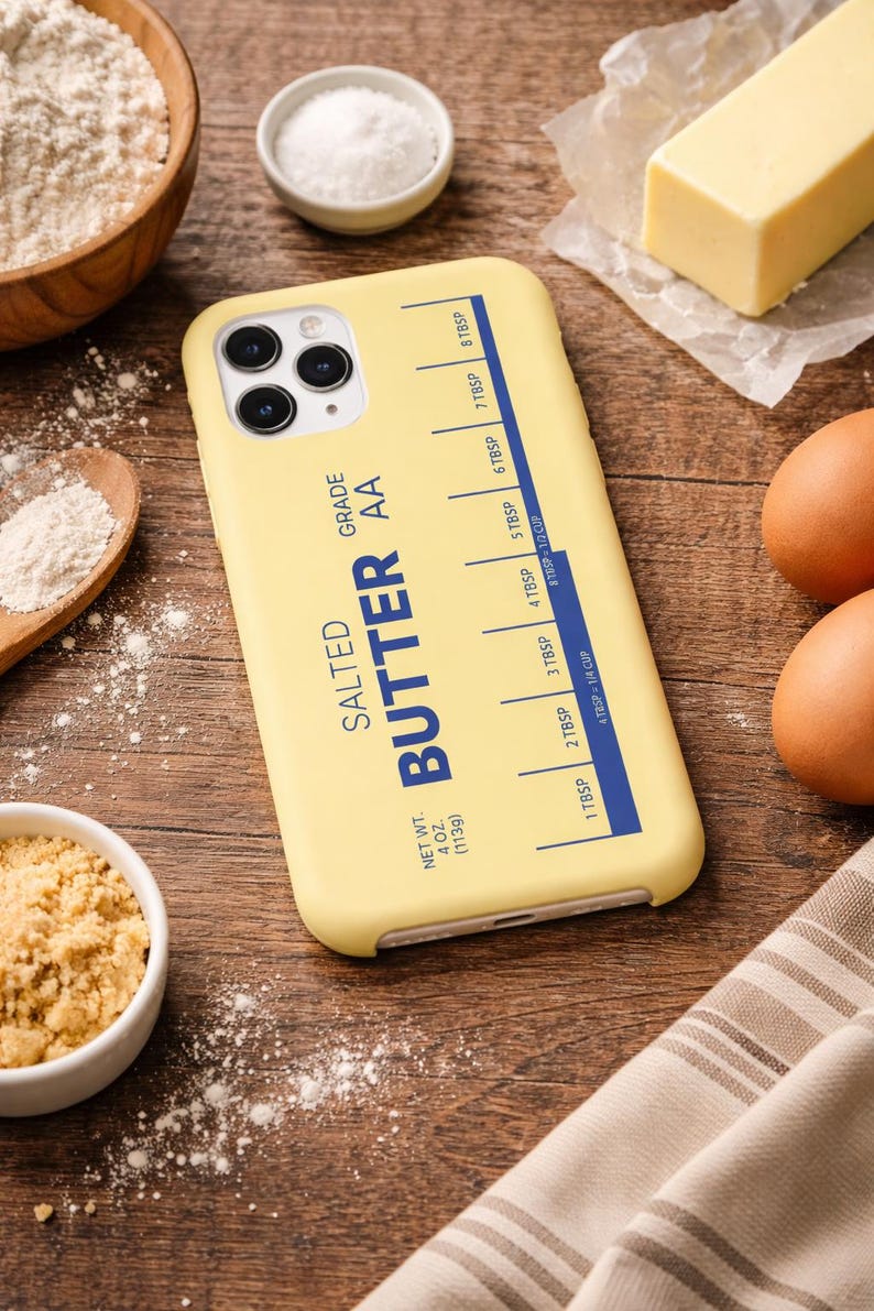 May include: A yellow phone case with a butter-themed design. The case features the words "SALTED BUTTER AA GRADE" and a measurement guide in tablespoons. Other baking ingredients like flour, eggs, and butter are in the background.