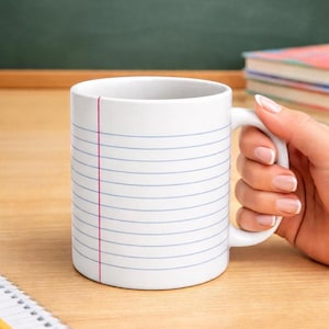 May include: White ceramic mug with a notebook paper design. The mug features blue horizontal lines and a red vertical line, resembling a sheet of lined paper. The mug is held by a person's hand, with a notebook and pencil in the foreground.