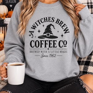 以下が含まれることがあります： A WITCHES BREW COFFEE CO、BREWED WITH A LITTLE MAGIC、Since 1962という黒いグラフィックプリントが施されたヘザーグレーのクルーネックトレーナー。魔女の帽子と小さな星のイラストが描かれています。