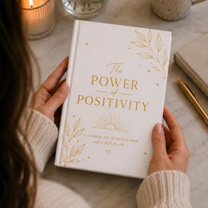 May include: A white hardcover journal with gold foil lettering that reads The POWER of POSITIVITY, A JOURNAL FOR A POSITIVE MIND AND A BETTER LIFE. The cover features gold line art of leaves, a sunburst, stars, and a small heart icon.