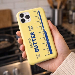 May include: A yellow phone case with a butter-themed design. The case features the words "SALTED BUTTER AA GRADE" and a measurement guide in blue. The case is held in a hand.