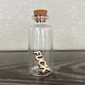 Pode incluir: Uma pequena garrafa de vidro com uma rolha de cortiça. A garrafa contém um recorte de madeira da palavra "FUCK".